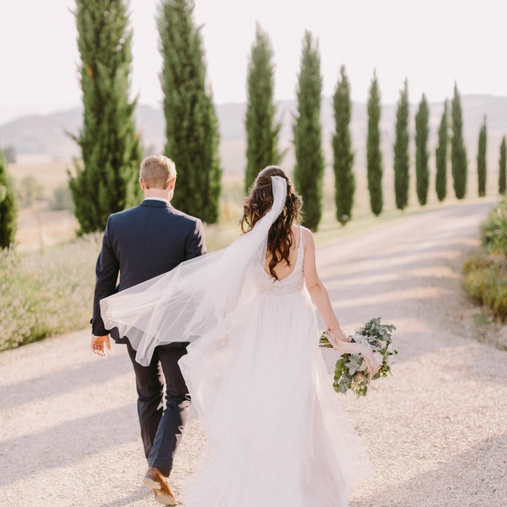 newly weds walking away from the camera in Tuscany wedding venue drive with cypresses on the sides and rolling hills on the background. the blonde groom is on the left and wears a blue suit and is holding the bride's left hand. The brunette bride has some loose curls that reach her shoulder blades, a veil and holding a loose bouquet on her right hand. her dress is simple and elegant white - sustainable luxury wedding