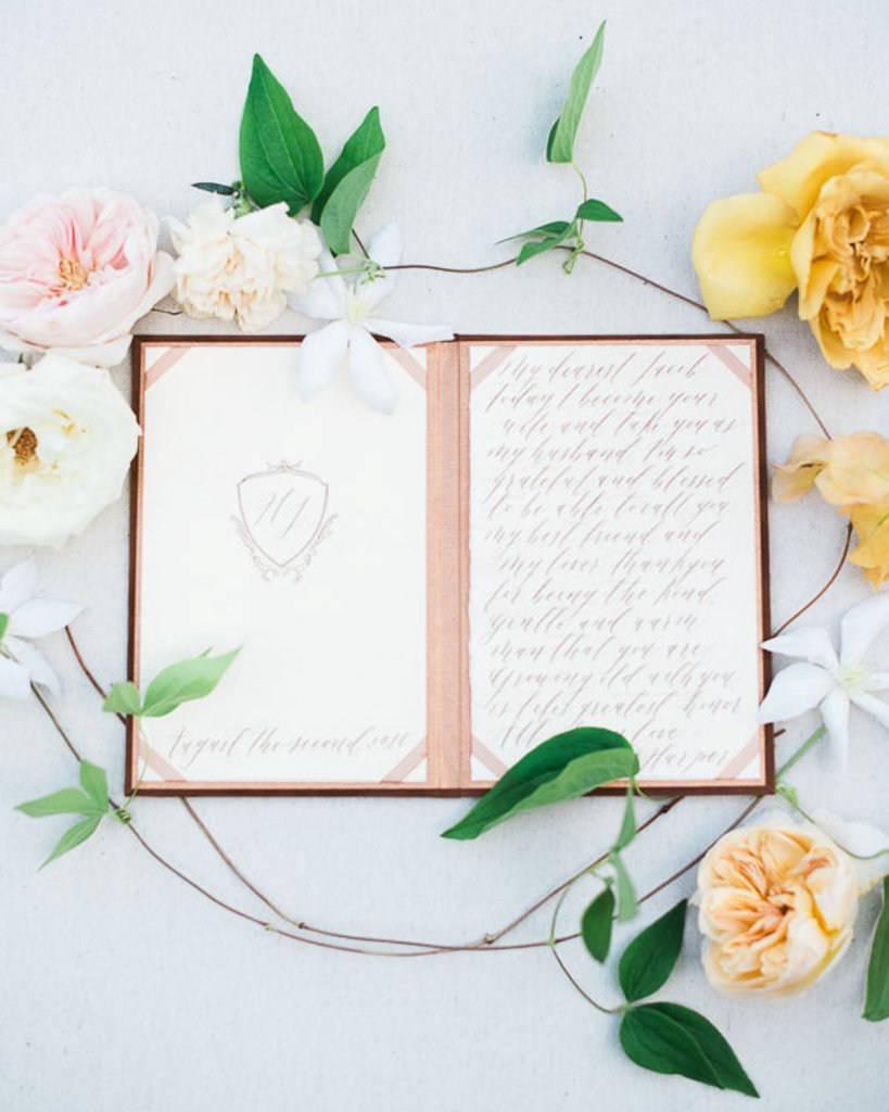 a flatlay of a fine art wedding guest book with blush decor and calligraphy, white roses and peach yellow peonies with some leaves wines