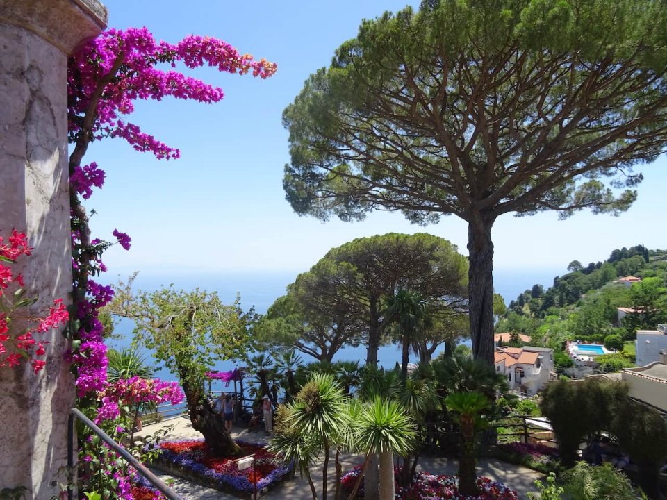Ravello seaside view from Villa Rugolo cliff and gardens, featuring a sea 2 pine trees in the centre-right, some pink and purple bougainvillae on the left hand wall, some red, purple floral garden un the garden  terrace underneath, green terraced hills on the right and a swimming pool - sustainable luxury wedding