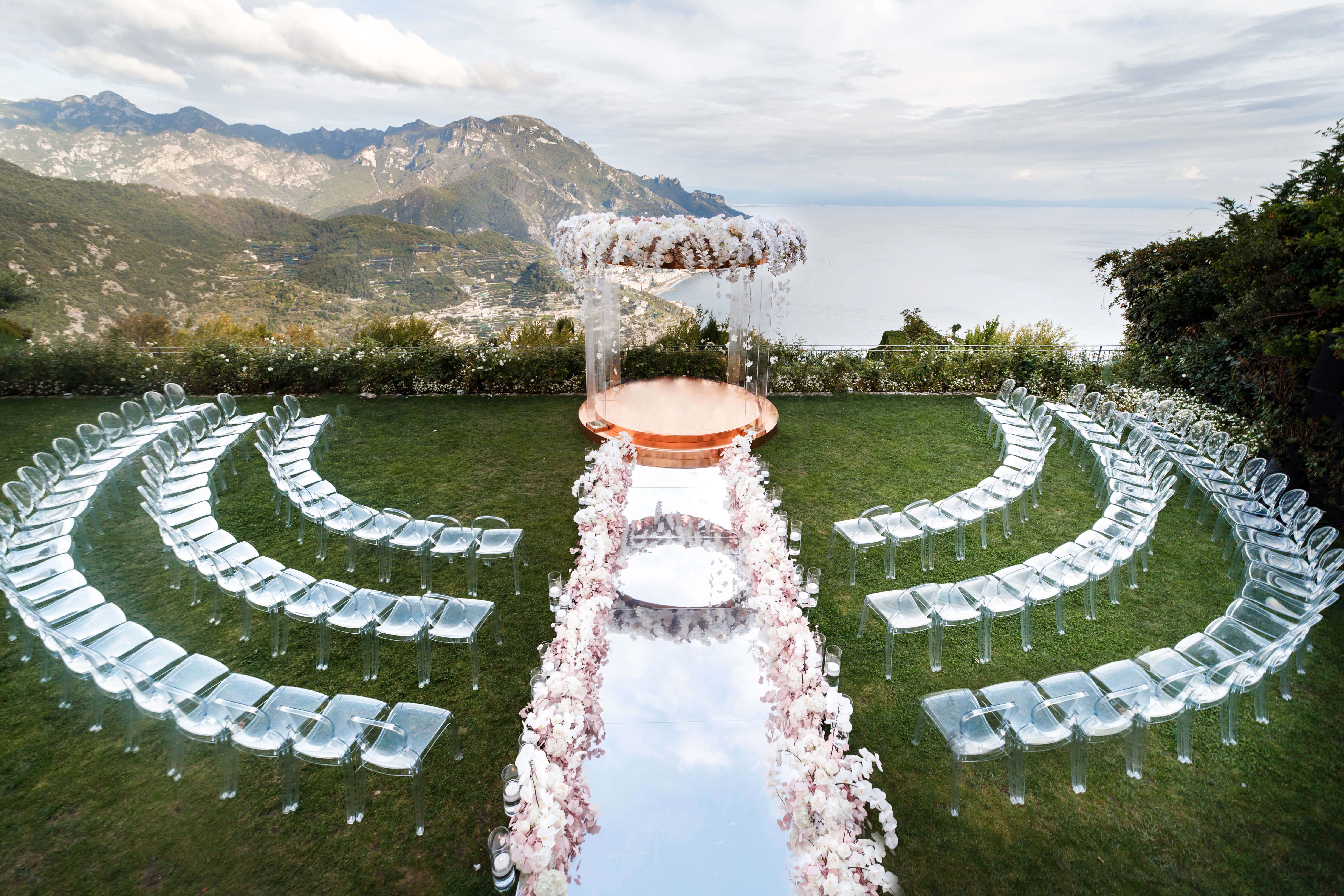 a clear glass chuppah with copper base and white floral decor on a lawn, overlooking amalfi coast by a cliff edge. there's a mirror aisle, low flral decor on the side of it and clear chairs with a semi circle ceremony set-up