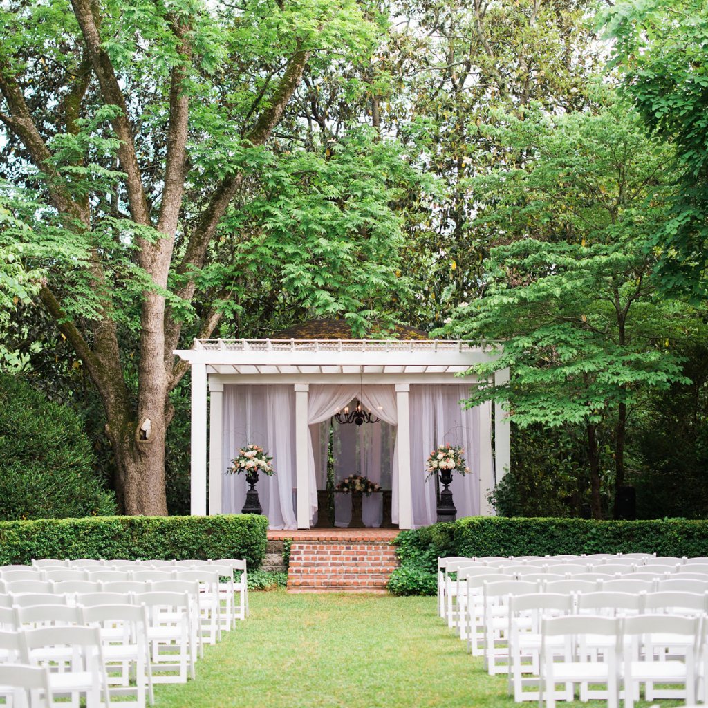 An outdoor wedding ceremony with tall trees as a background, a small elevated white wooden chuppah with white fabric hanging on all sides, featuring 2 black high flower vases on both sides, 4 red brick steps comin down the green grass aisle, white plain wooden chairs on both sides - sustainable luxury wedding