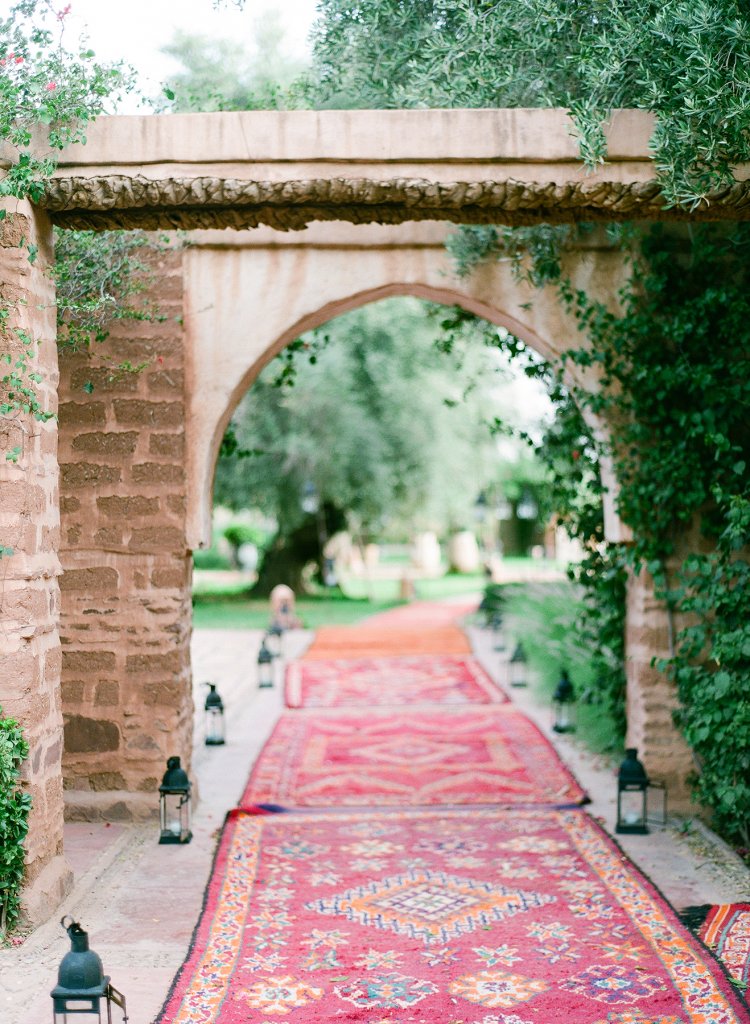 Red Persian carpets laid on a garden  path outdoors, a red brick arch, with black metal lanterns on both sides, leading to a wedding reception - sustainable luxury wedding