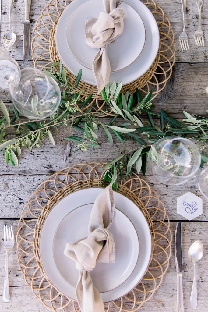sustainable wedding table decor rentals featuring a naked table olive branches runner, wick chargers, sand napkin on a white plate, wine glass, personalised marble place card and silver cutlery - sustainable luxury wedding
