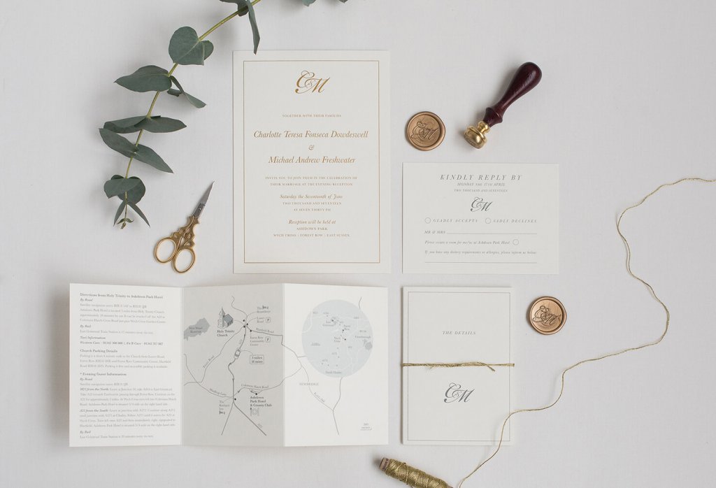 a flatlay of cream Wedding stationary suite with gold letterpress, sage leaves, wax stamp in dark wood and brass, small brass scissors RSVP cards, copper wax seals, and a small map