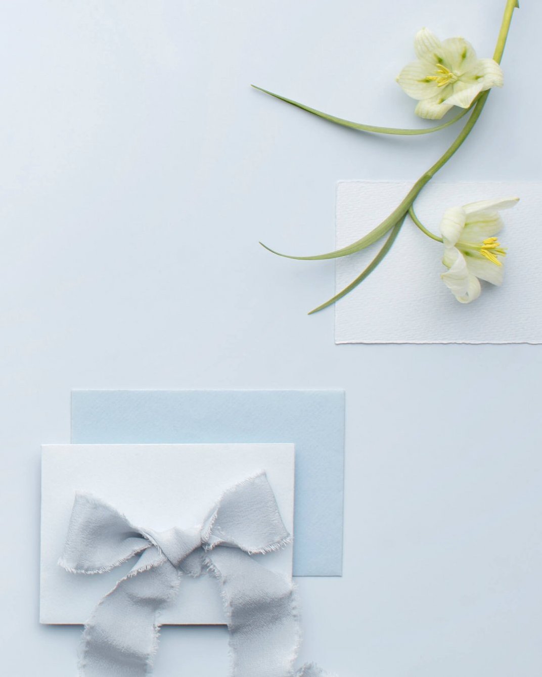 baby blue wedding invite envelope and white invite with a silky baby blue ribbon on top, laying on a baby blue background and some white and yellow stemmed flower on the top right hand corner