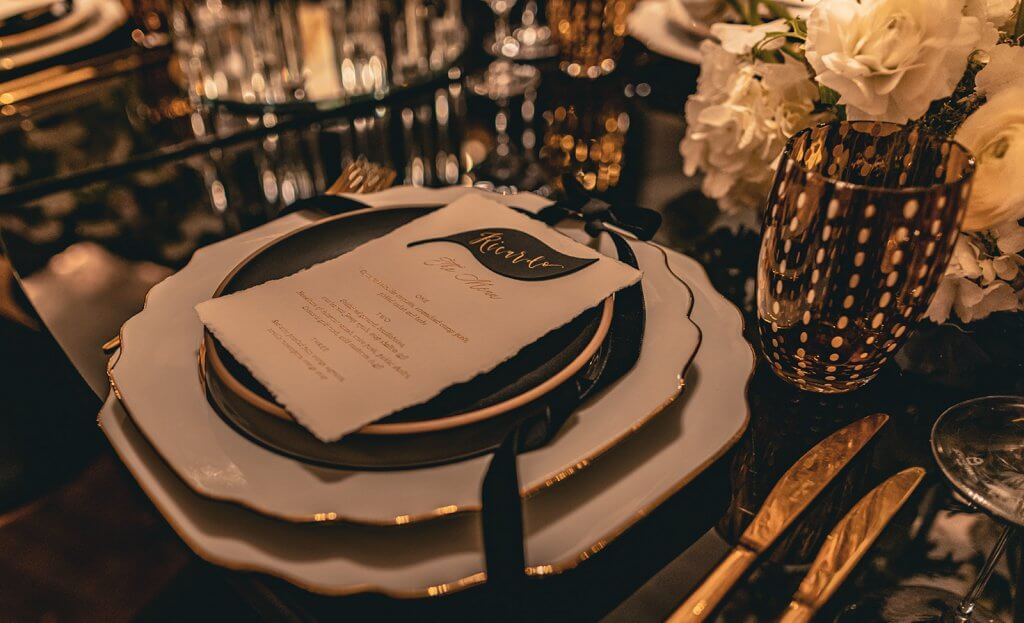 gold rimmed scalloped white plate set with white menu and black velvet ribbon on a glossy black serpentine table, amber water speckled glass and gold cutlery