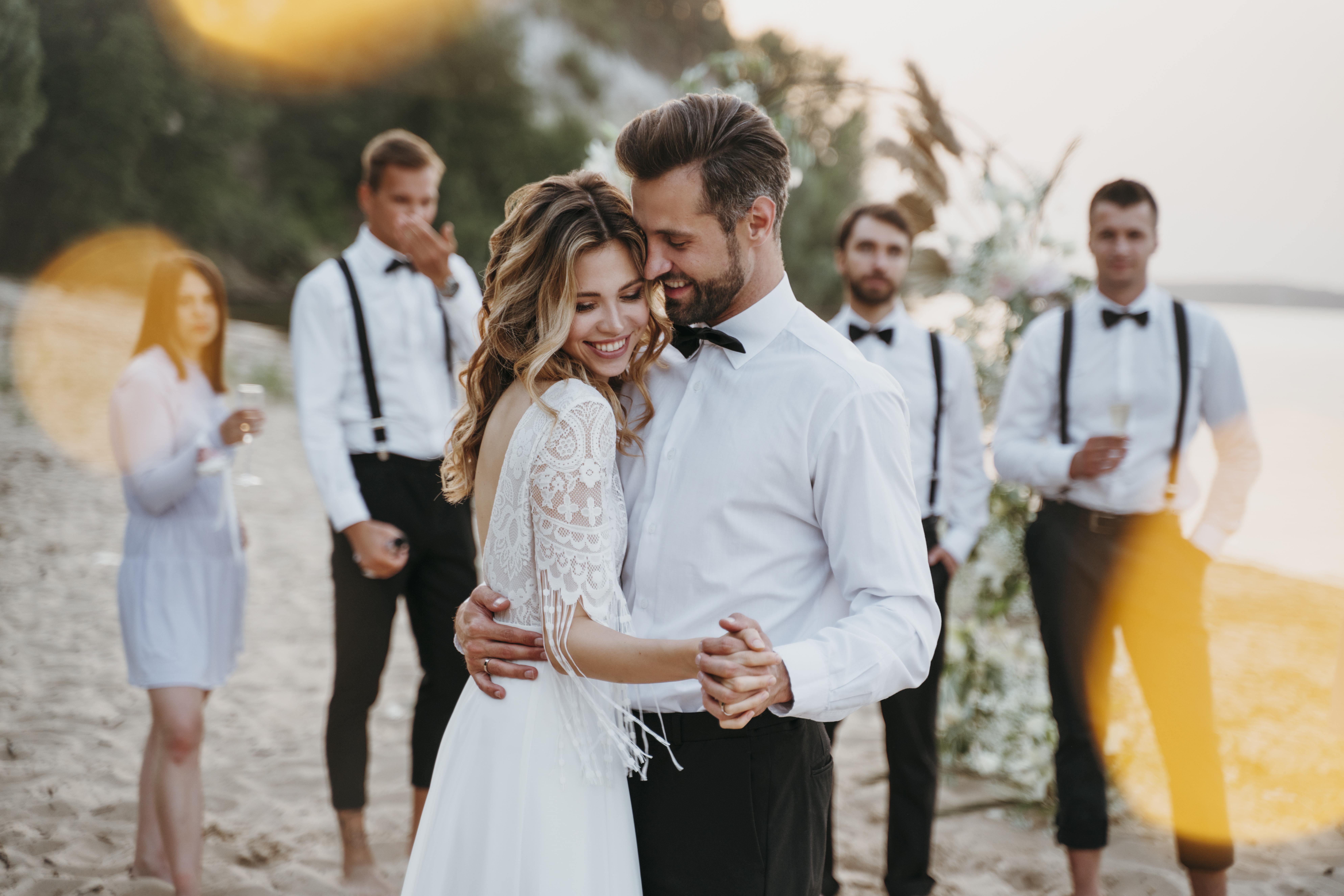 Bride and groom hugging while listening to best man wedding speech with their groomsmen in the background toasting to the newlyweds during sunset
