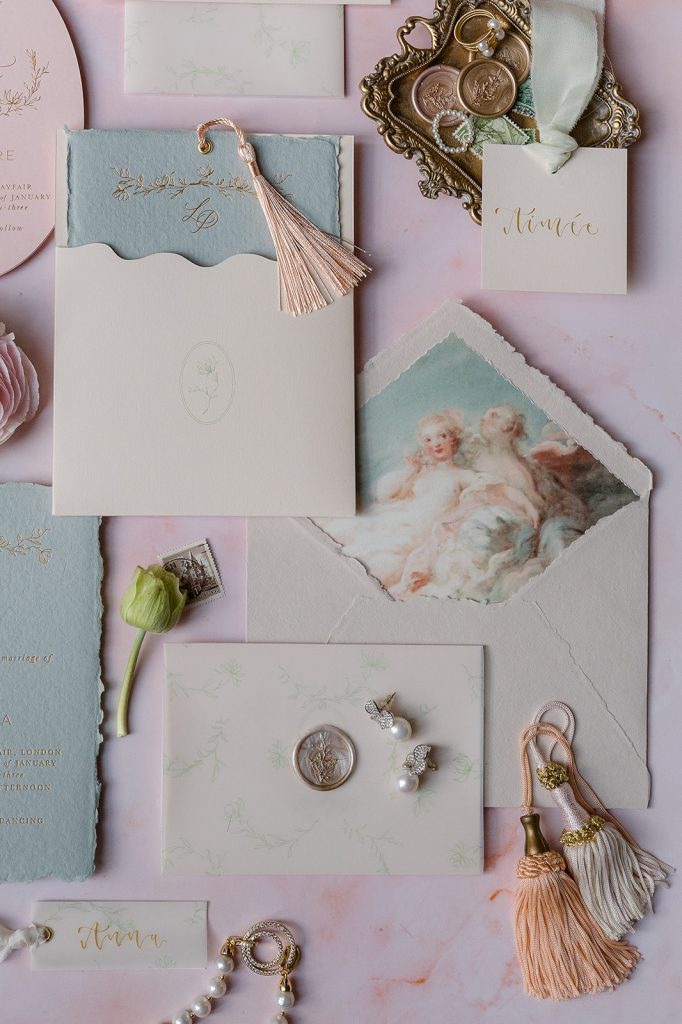 flatlay of Pink save the dates, jade wedding invite and cream envelopes with a bespoke lining representing dartmouth house ceiling cherubs' frescoe Stationary suite with gold calligraphy and letterprint, letterpress writing on a pink backdrop, with some peach, cream and gold tassels