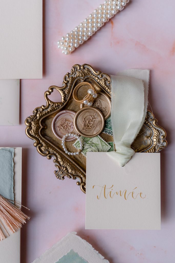 flatlay close-up of small brass trinket trat with gold and copper wax seals, a cream place card with gold calligraphy and aqua silk ribbon, a pearl bracelet