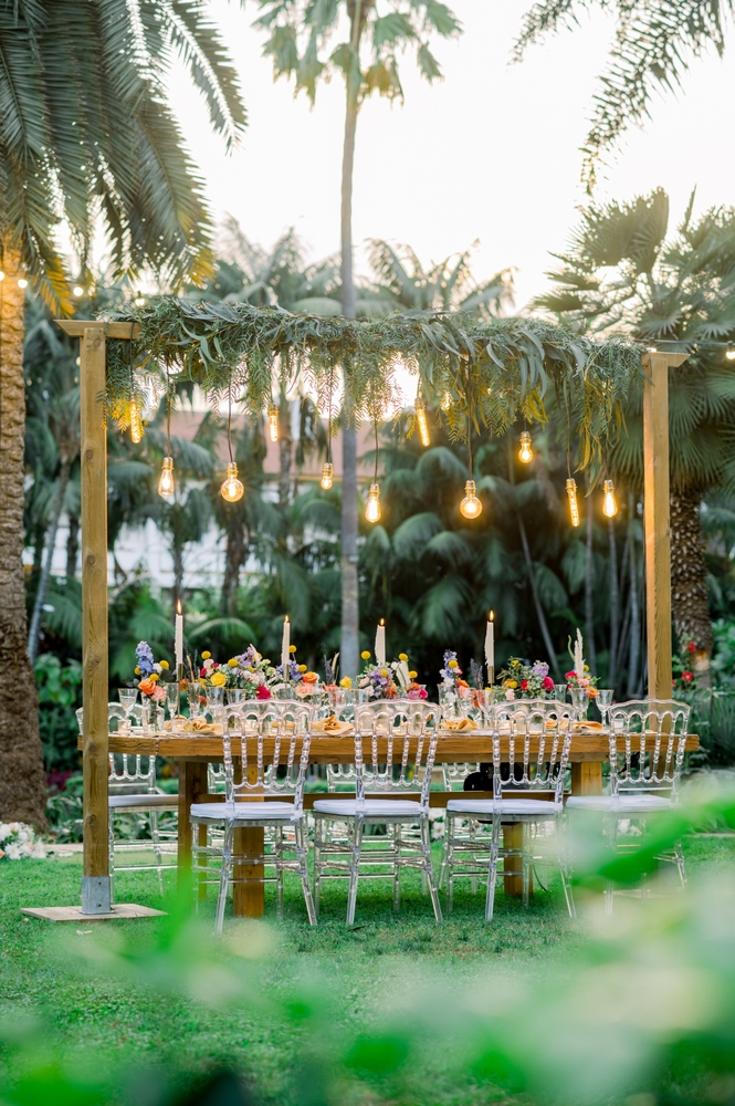 a welcome party table with dtring bulb lihts over the length of the table, with napoleon ghost chairs, and red, blue and yellow flowers, on a lawan with palms on the background