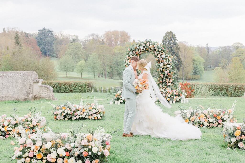 bride and groom kissing standing in front Paris Wedding Chateau garden wedding ceremony set up with Pastel Floral arch and meadows in an S shape on a lawn in front of Chateau Bouffemont