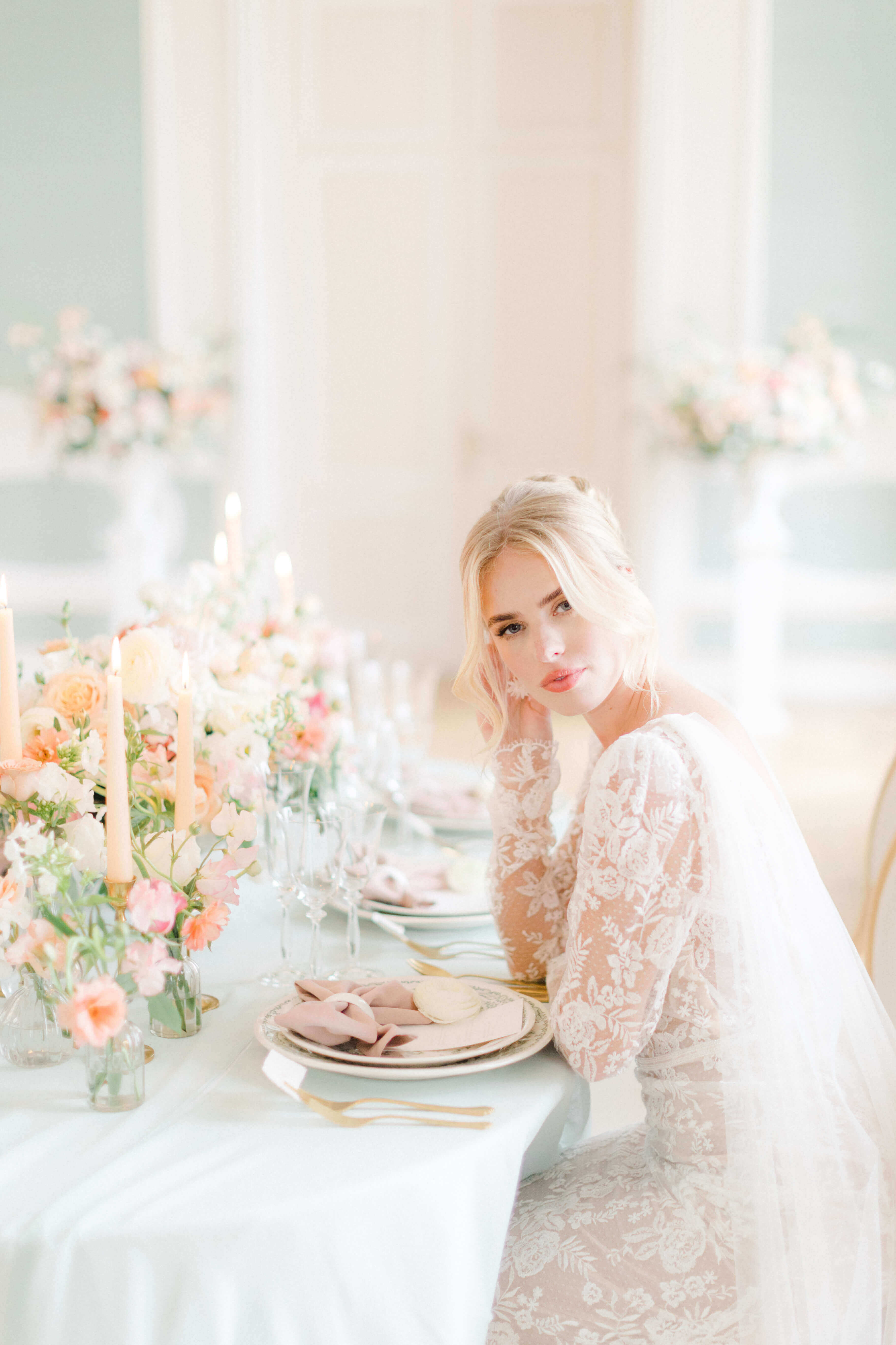 Bride sitting at pastel wedding reception table. The bride's hair is tied in a high bun and she's wearing a low cut decoletté lace dress with long sleeves. the table linen is mint and there are crystal glassware, white and green porcelain tableware, golden cutlery, pink, peach, cream pastel hues of floral centrepieces and light peach tapered candles.