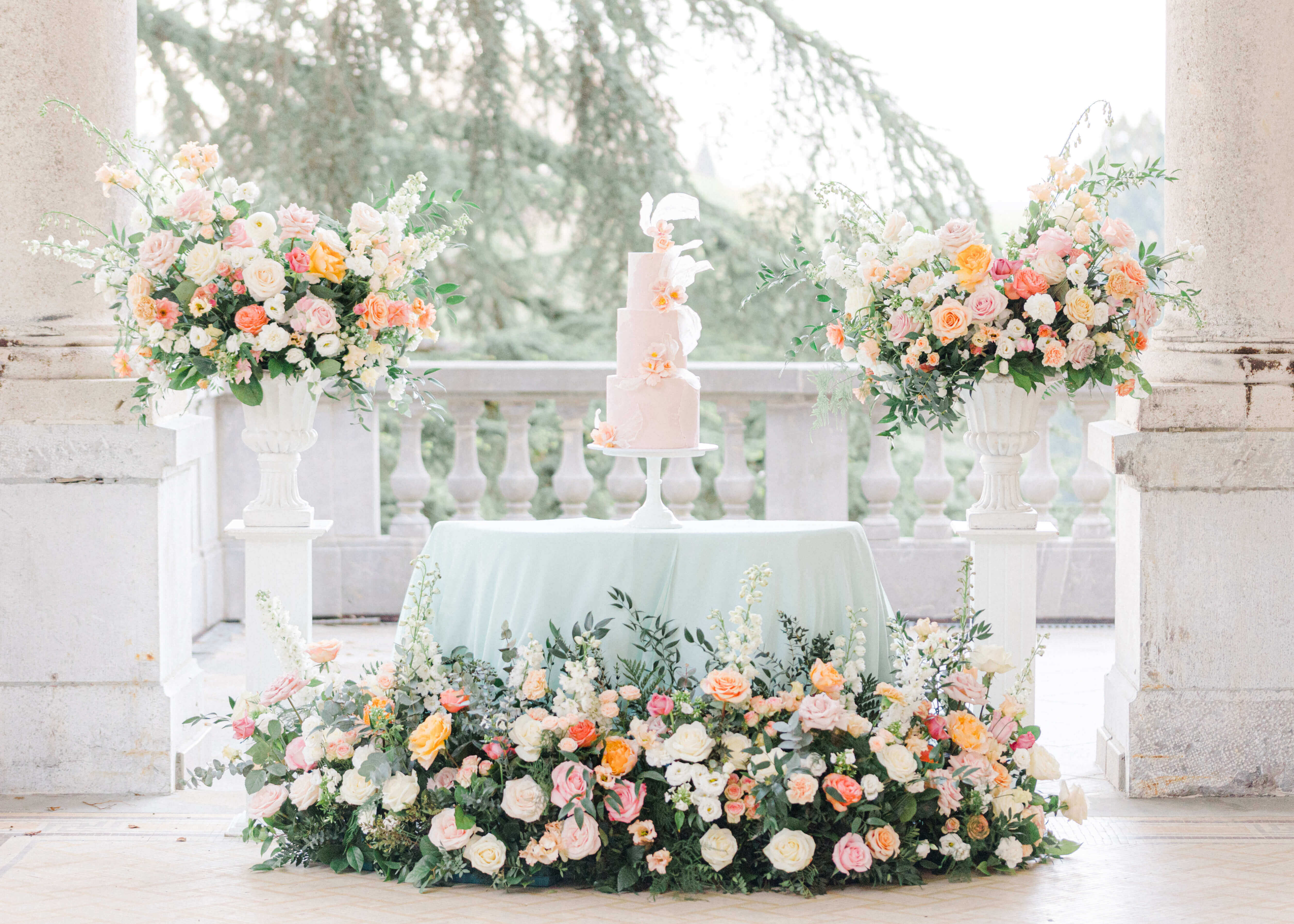  pastel peach cake under a terrace at chateau bouffemont.  the cake is sitting on a table covered by mint linen, surrounded by pastel pink, peach and cream floral meadows and and on both sides 2 plinths with similar florals