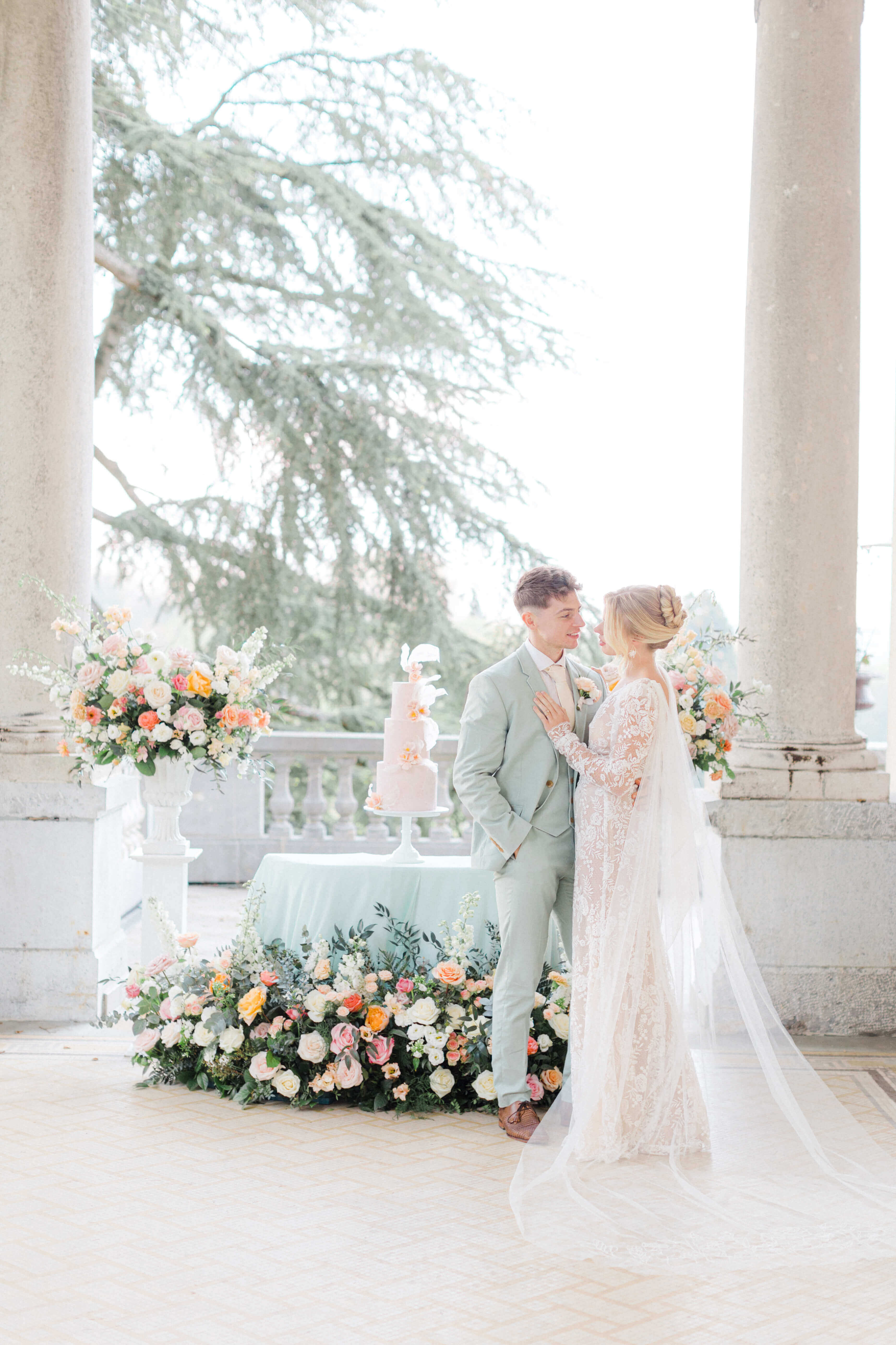 pastel peach cake under a terrace at chateau bouffemont.  the cake is sitting on a table covered by mint linen, surrounded by pastel pink, peach and cream floral meadows and and on both sides 2 plinths with similar florals. Newly weds in front of it. the groom is wearing a mint tux and cream tie on a white shirt. The bride's hair is tied in a high bun and she's wearing a low cut decoletté lace dress with long sleeves