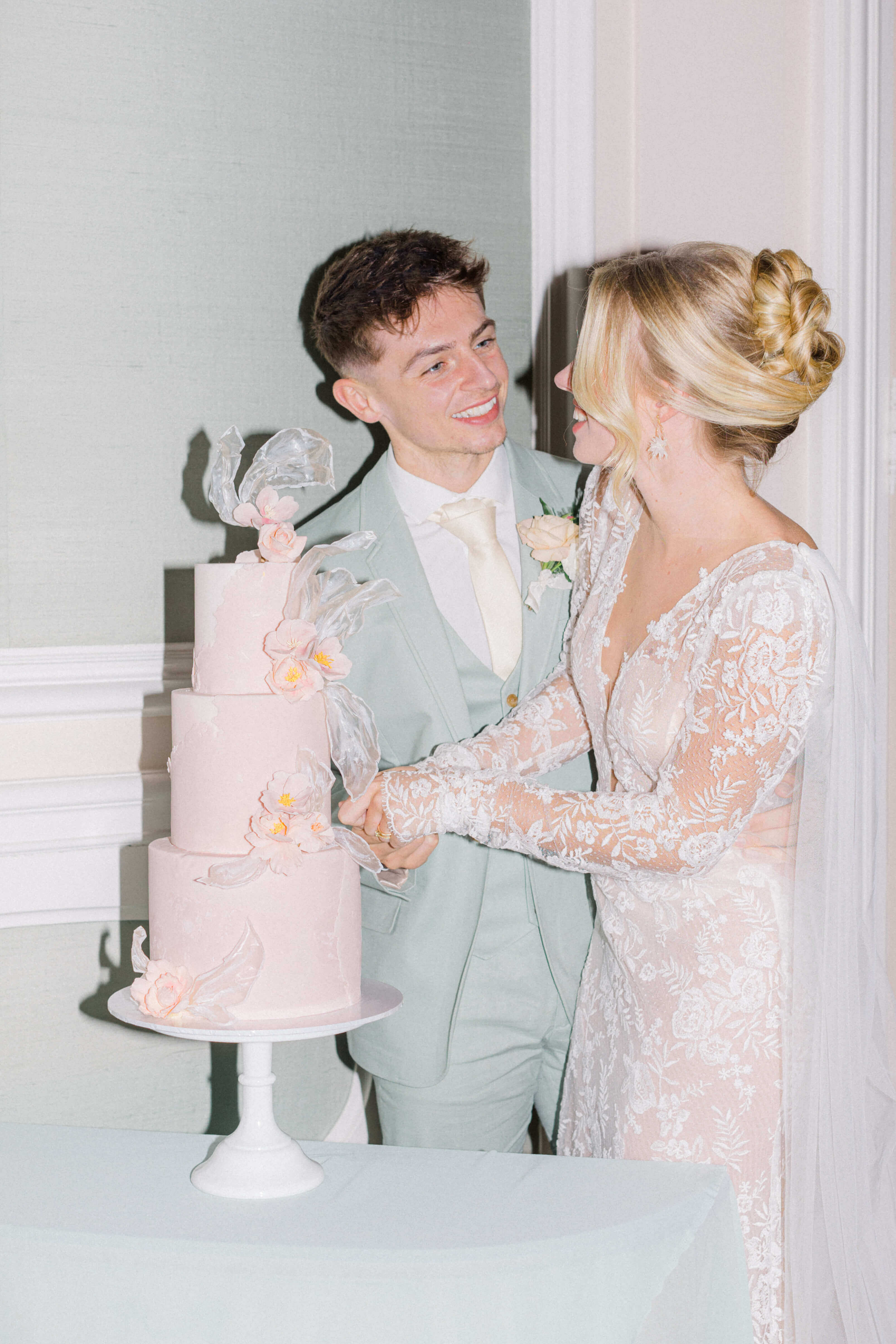 groom and bride cutting pastel peach cake looking at each other. the groom is wearing a mint tux and cream tie on a white shirt. The bride's hair is tied in a high bun and she's wearing a low cut decoletté lace dress with long sleeves