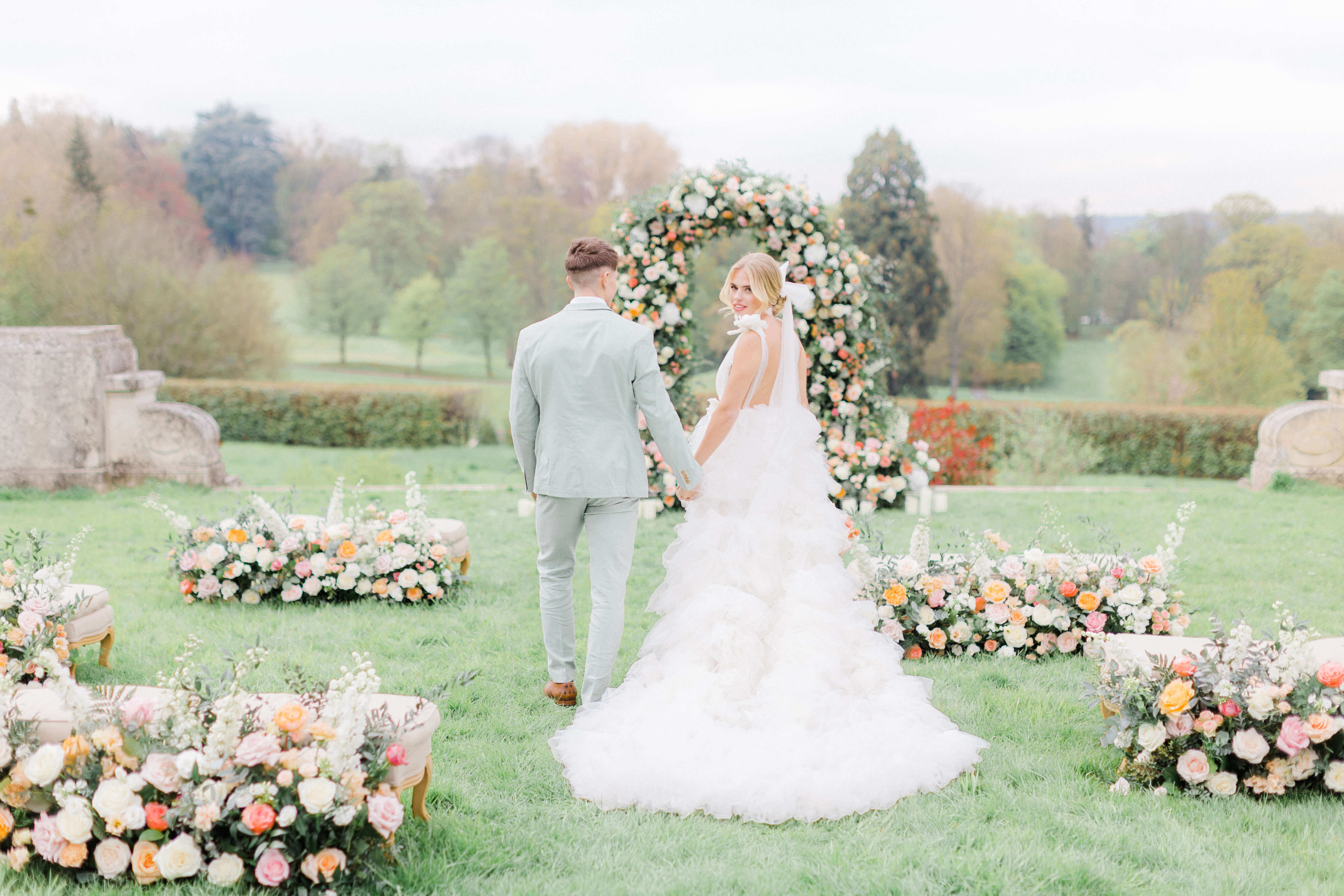 bride and groom walking towards a wedding ceremony pastel floral arch and meadows in an S shape on a lawn in front of Chateau Bouffemont