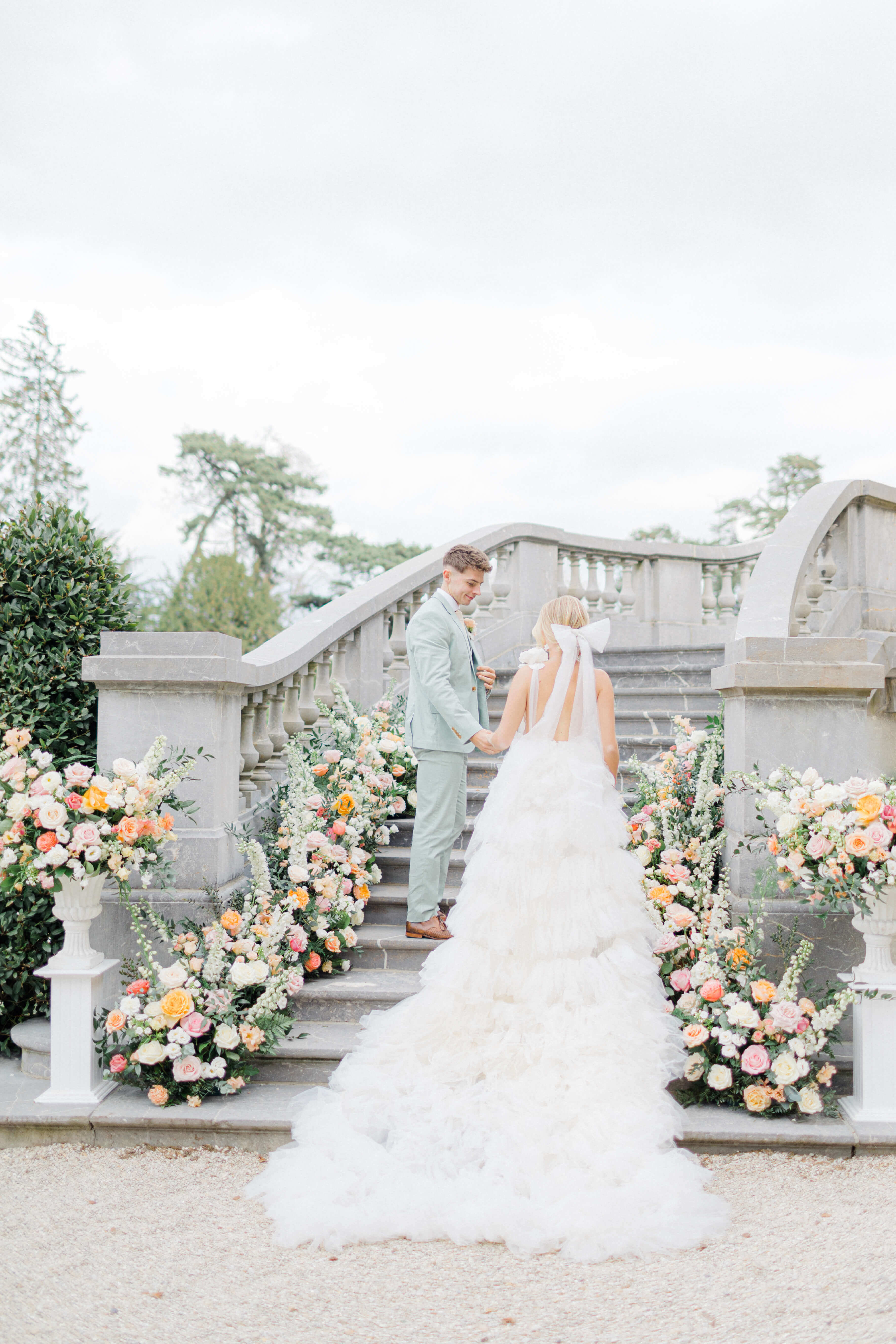 Pastel floral meadows in a semi arch on a staircase at chateau bouffemont, with newly weds walking towards it. The groom is wearing a mint tux and cream tie on a white shirt. The bride's hair is tied in a high bun and she's wearing a low cut decoletté ruffle white dress.