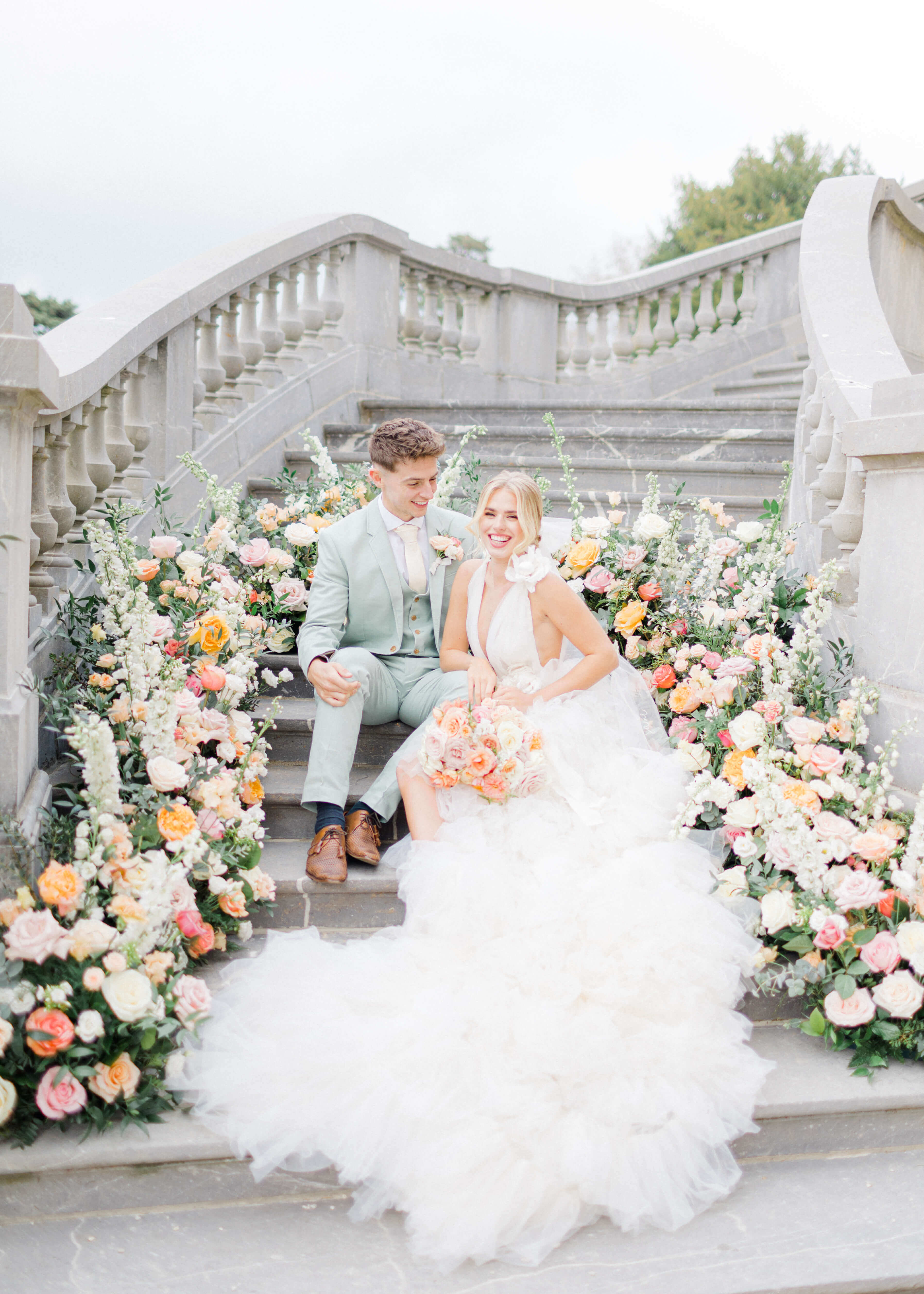 Pastel floral meadows in a semi arch on a staircase at chateau bouffemont, with newly weds sitting  in front of it. The groom is wearing a mint tux and cream tie on a white shirt. The bride's hair is tied in a high bun and she's wearing a low cut decoletté ruffle white dress.