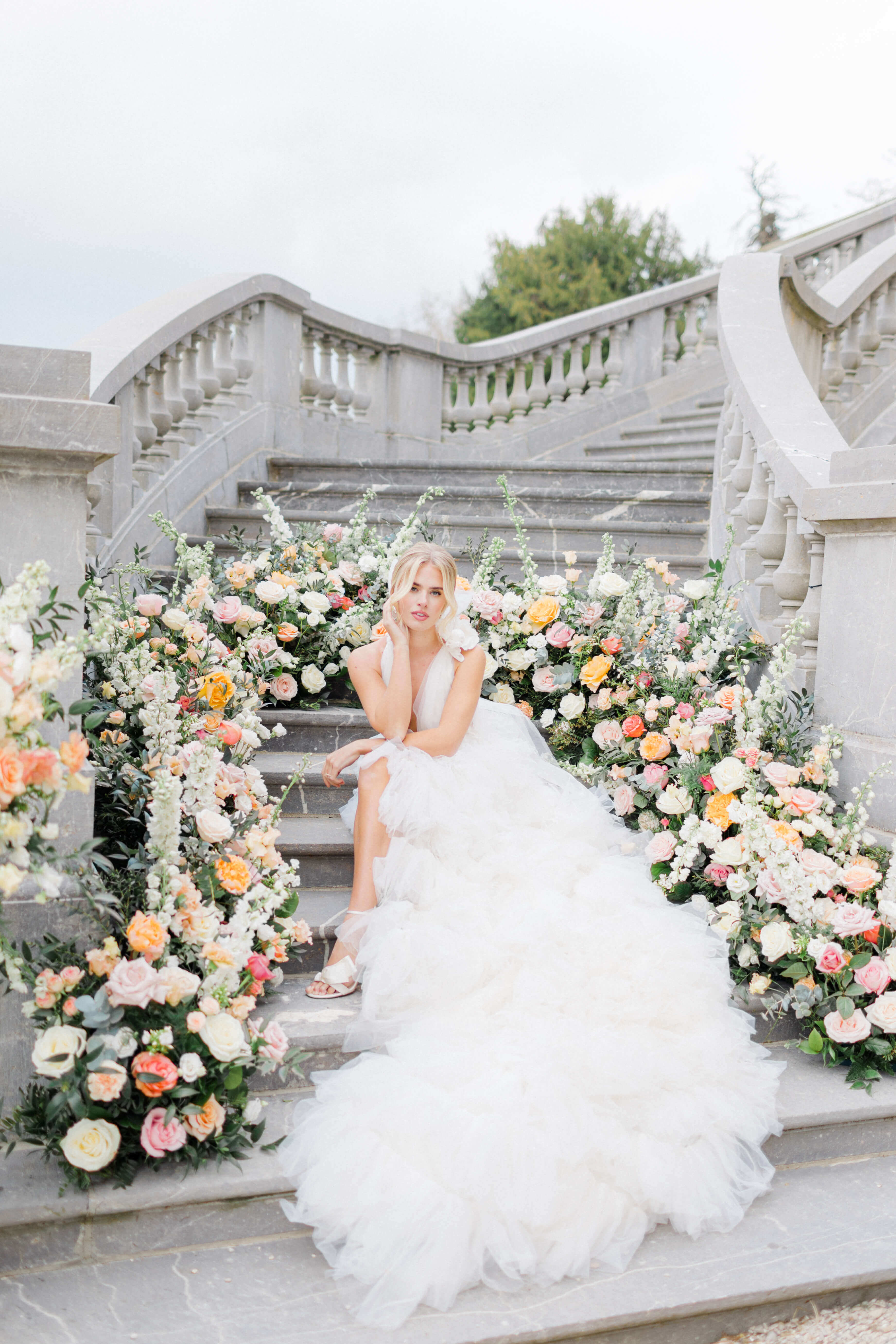 Pastel floral meadows in a semi arch on a staircase at chateau bouffemont, in front of which a bride is sitting with her ruffle white dress, 