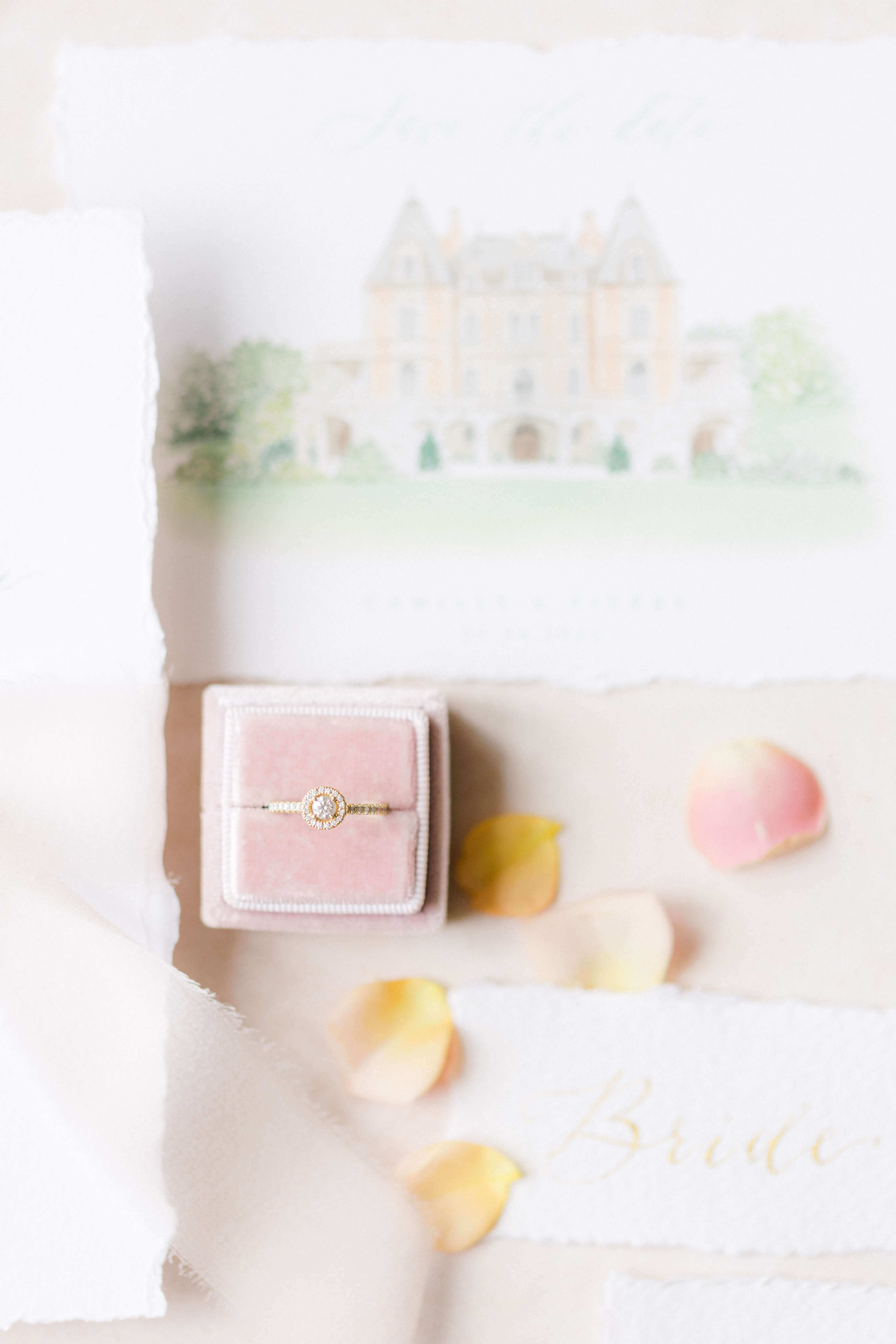  pastel pink ring box holding a diamond engagement ring, sitting on a wedding invitation representing Chateau Bouffemont, some pink and peach rose petals scattered and a white placecard with Bride written on it