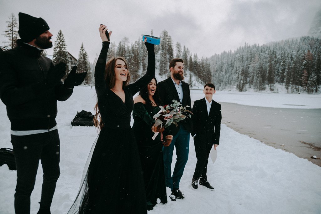 bridal party dresses in black a snowy lake ceremony, bridesmaid holding a bouquet and another one throwing hands in the air as ceebration