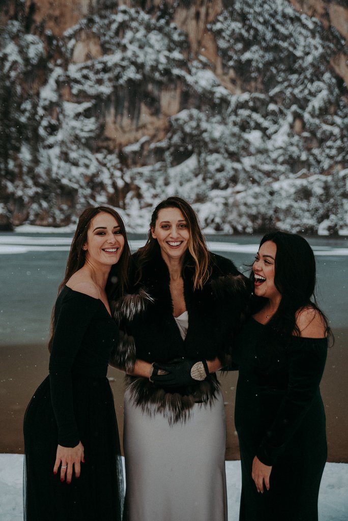 bride with black furry shawl and bridesmaids  party dresses in black a snowy lake ceremony, bridesmaid holding a bouquet