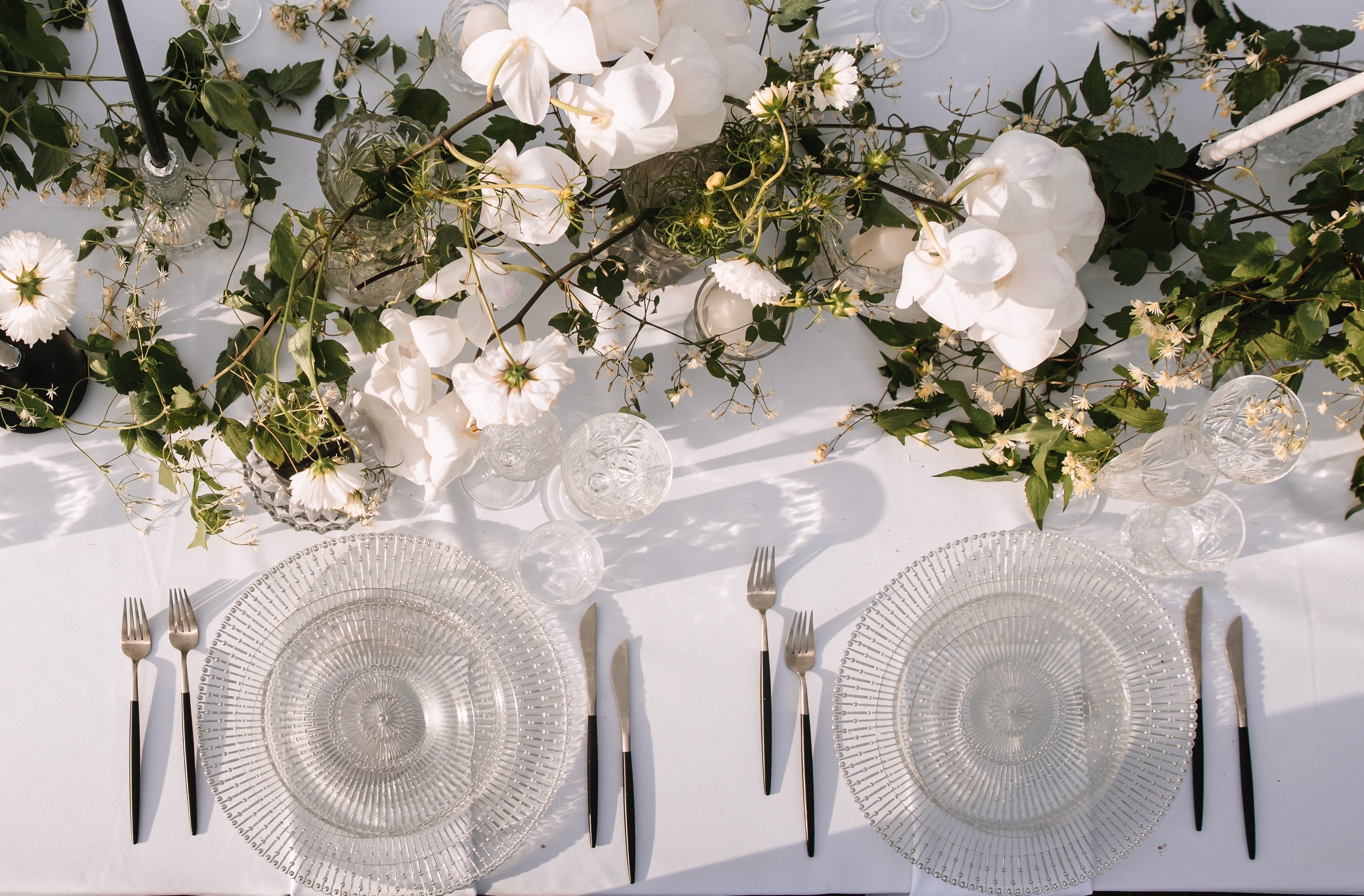 a tabletop with white linen cloth, clear glass embellished chargers and glasses, white and green floral runner and silver and black modern cutley
