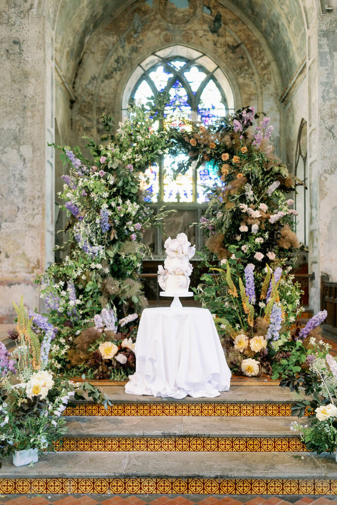 powder blue, lilac and copper palette floral arch on steps of a former church altar, matching the stained window glass colours. The white and lilac iris wedding cake is displayed in front of the arch, on a round cake tabled covered with white linen 