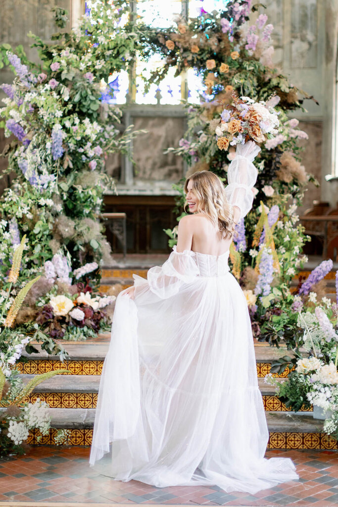bride portrait from the back holding her skirt with her left hand and holding up her bouquet with her right one smiling. In the background Wedding ceremony with lilac, powder blue and copper floral arch and meadows on the sides