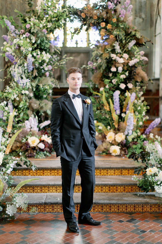 blonde groom in black tux, posing with hands in pockets in front of Wedding ceremony with lilac, powder blue and copper floral arch and meadows on the sides