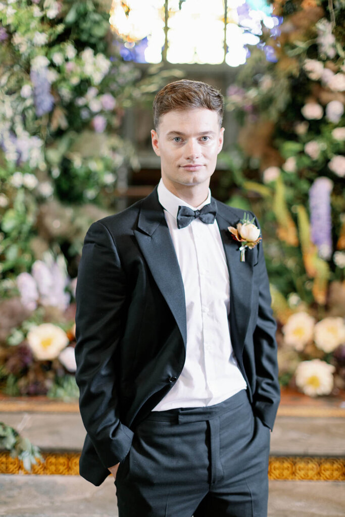 Groom's portrait smiling at the camera. he's wearing black tux with bow tie and hands in his pockets Behind him In the background Wedding ceremony with lilac, powder blue and copper floral arch and meadows on the sides