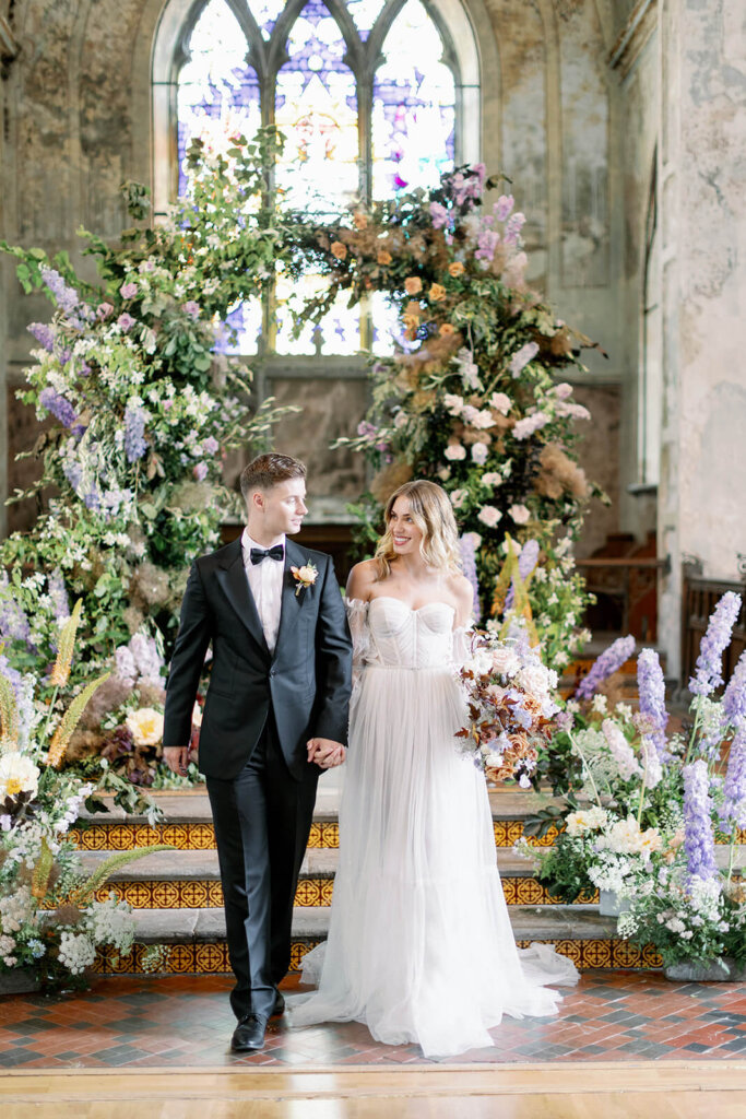blond groom on the left hand in hand with blonde bride, walking away from In the background Wedding ceremony with lilac, powder blue and copper floral arch and meadows on the sides