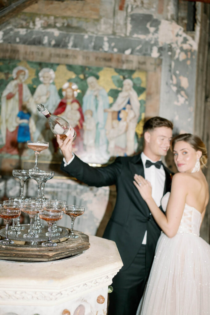bride and groom behind a champagne tower set on a baptism fountain in a former church, while groom is pouring pink champagne. behind them a painting of the last supper. she's looking at the camera and he looks at her