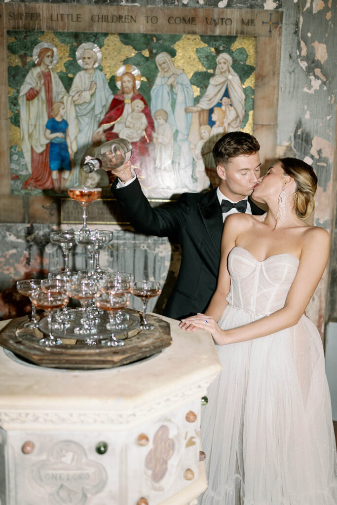 bride and groom kissing behind a champagne tower set on a baptism fountain in a former church, while groom is pouring pink champagne. behind them a painting of the last supper
