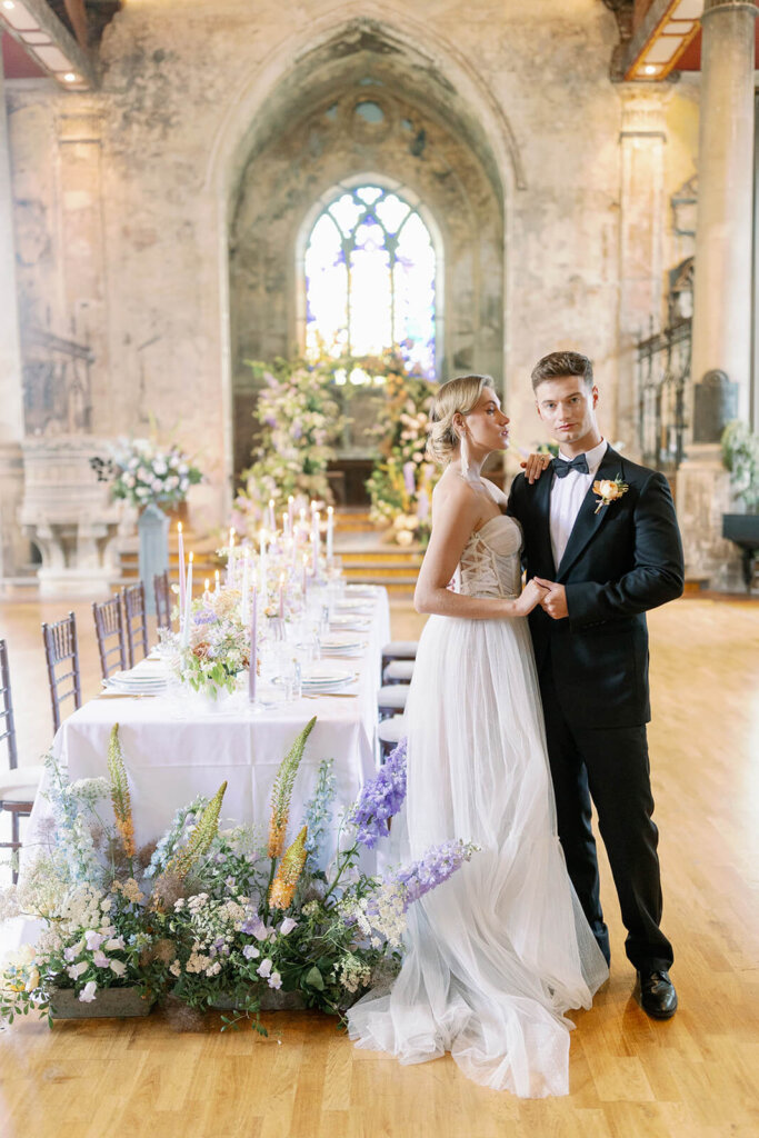 long wedding table with floral meadows at the front, floral arch iand a cake in the background under a a church window. a blonde groom in black tux is walking in front of it and holding his blonde bride's hand, and he's looking at the camera