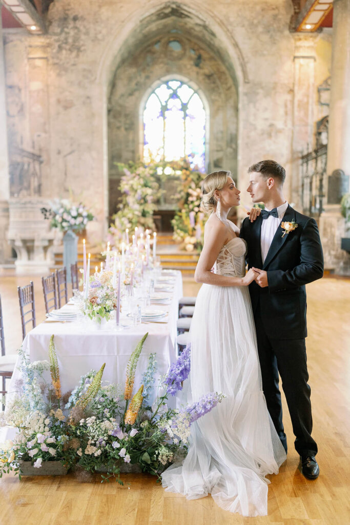 long wedding table with floral meadows at the front, floral arch iand a cake in the background under a a church window. a blonde groom in black tux is walking in front of it and holding his blonde bride's hand, 