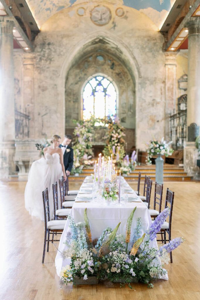 long wedding table with floral meadows at the front, floral arch iand a cake in the background under a a church window. a blonde groom in black tux is walking behind it and behind his blonde bride's hand, who's checking the table details