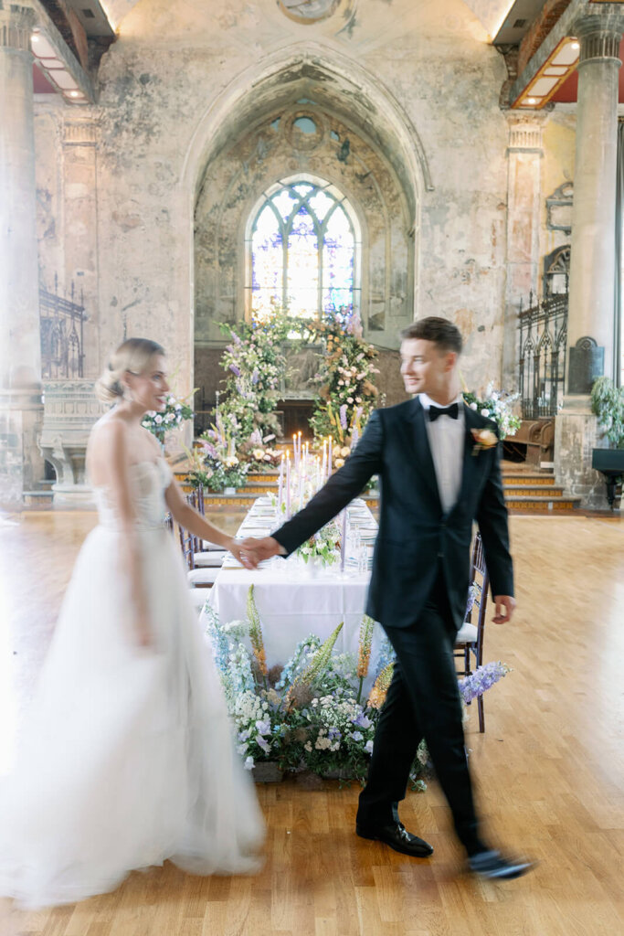 long wedding table with floral meadows at the front, floral arch iand a cake in the background under a a church window. a blonde groom in black tux is walking in front of it and pulling his blonde bride's hand, who's walking  behind him
