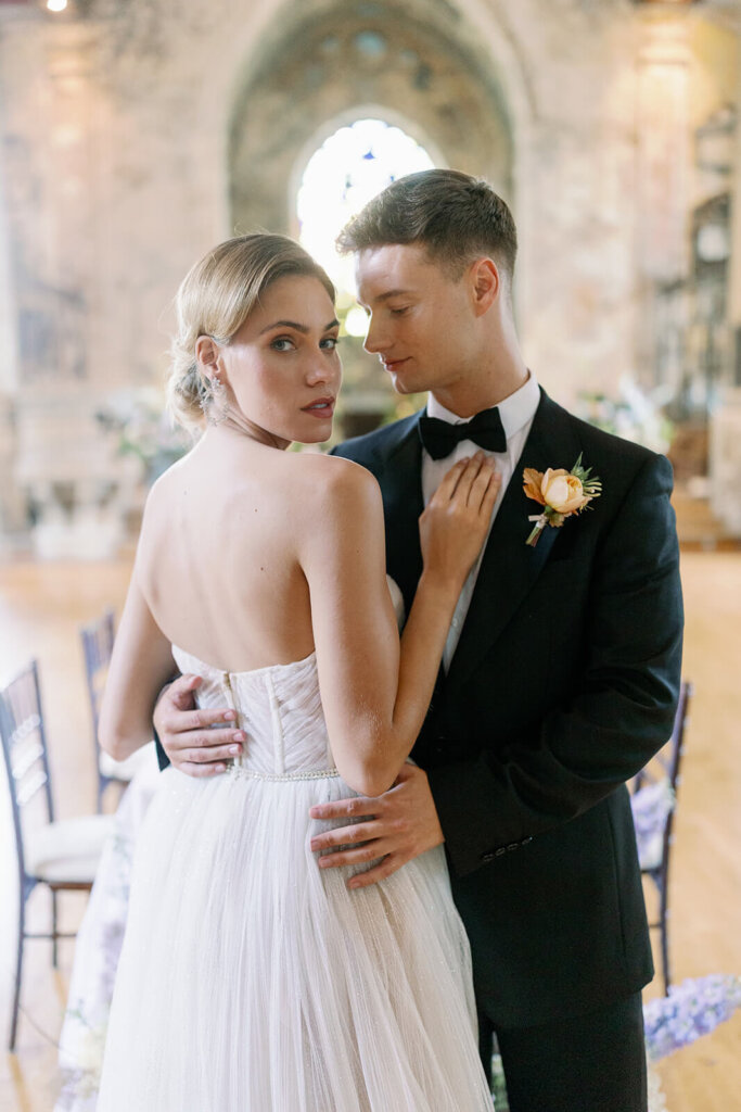 a blonde groom in black tux posing in front of his bride, who's staring at the camera, while his looking at herin front of it and pulling his blonde bride's hand, who's walking  behind him