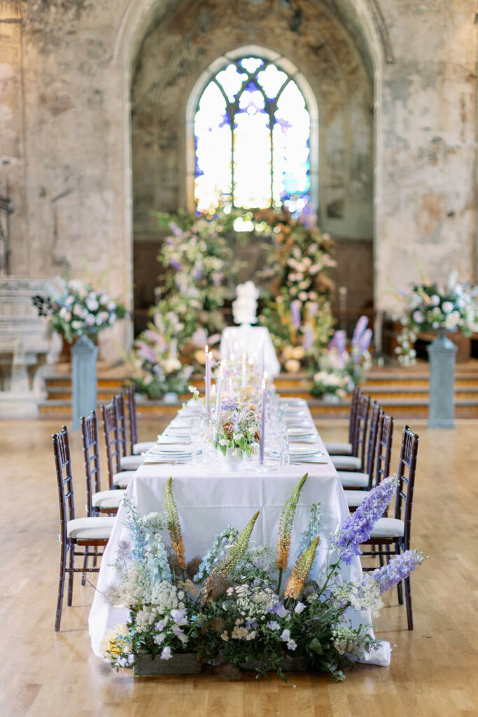 long wedding table with floral meadows at the front, lilac, powder blue and copper floral arch iand a cake in the background under a a church window