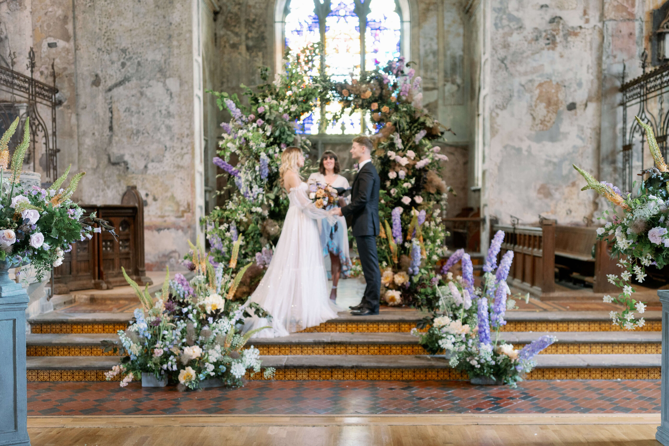 Wedding ceremony with lilac, powder blue and copper floral arch and meadows on the sides, bride on the left, groom on the right and celebrant behind them. A stained glass windo in the same colour palette behind the arch