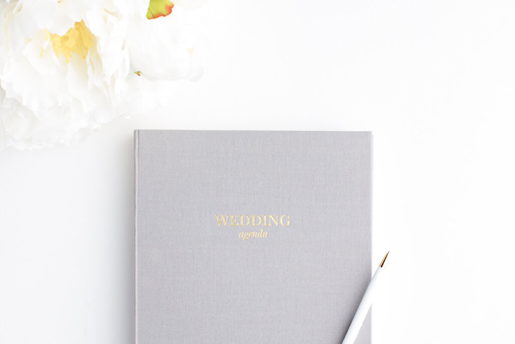 a silver wedding planning agenda cover with golden letters, a white pen, and white and yellow peonie on the left hand corner