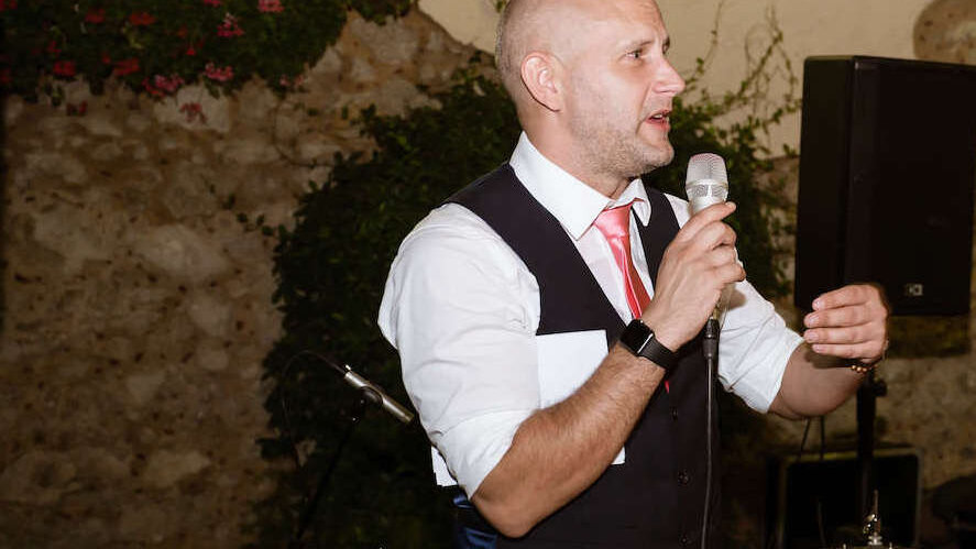 best man holding a microphone during his wedding speech addressing the audience. In the background drums from the wedding banc