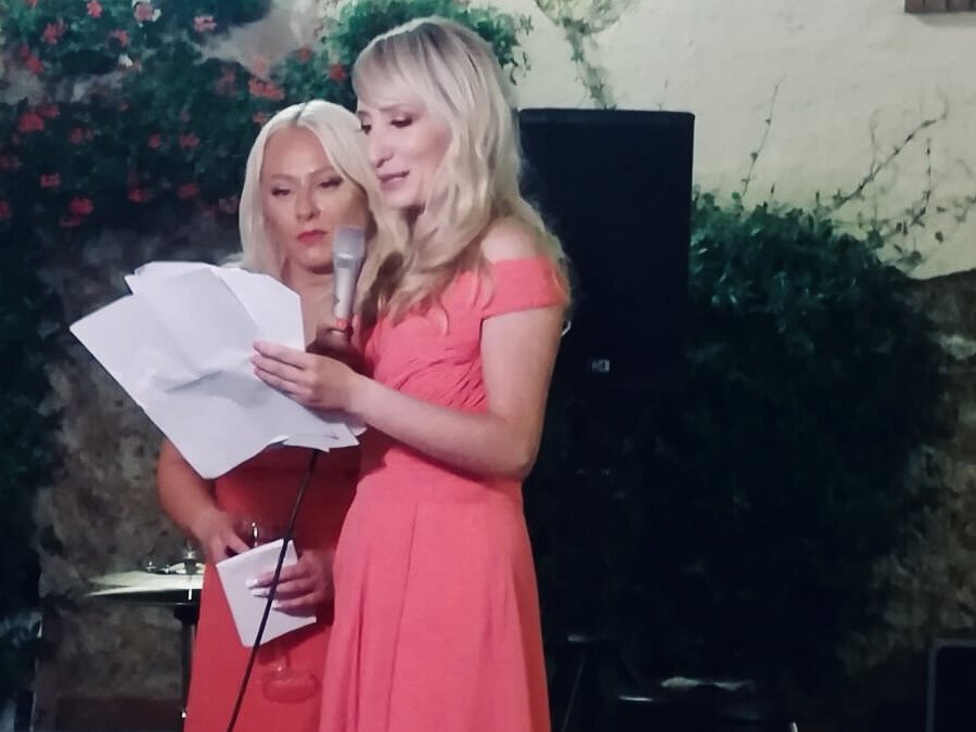 2 blonde bridesmaids reading their a Wedding Speech holding a microphone and paper sheets, wearing a coral gown at an outdoor wedding in italy 